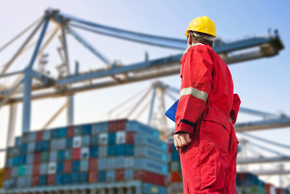 Worker in red overalls and yellow hard hat stands in a shipping yard, holding a blue clipboard